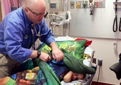 Tae hides under the covers during treatment as Duncan Stearns, MD, Director, Pediatric Neuro-Oncology, Angie’s Institute at UH Rainbow, uncovers and checks on him.